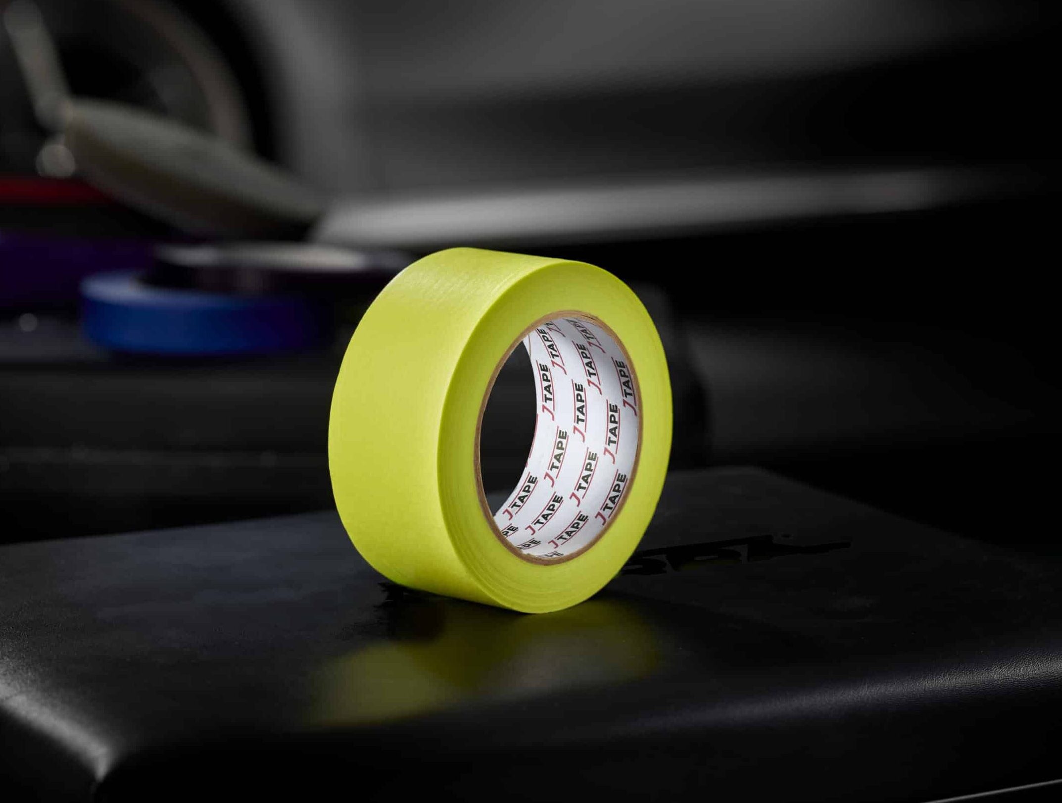 A roll of bright yellow wide tape, standing upright on a dark surface, with a blurred background that includes another roll of tape and a car door.
